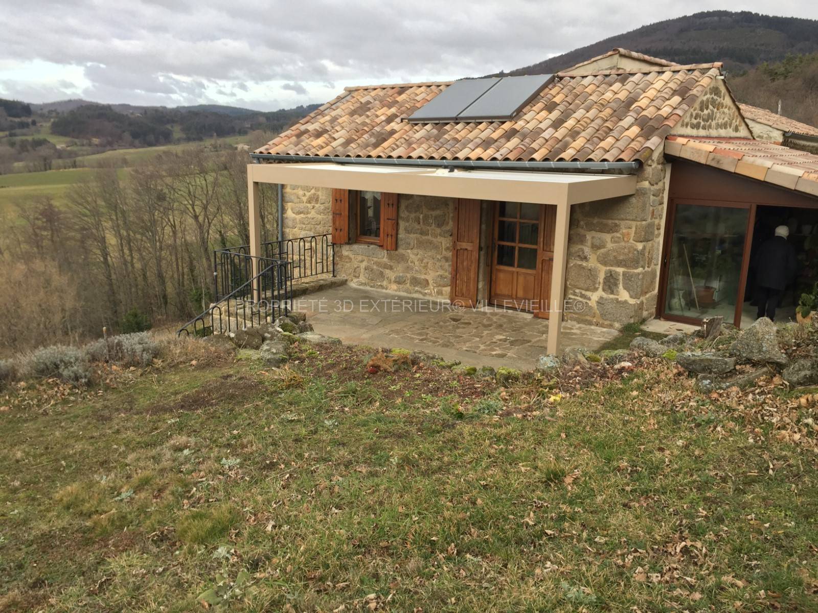 Réalisation d’une pergola bioclimatique couleur standard (Ral1019) beige gris et lames orientables blanches orientables adossée sur un mur en pierres d’une maison située à Châteauneuf de Vernoux dans